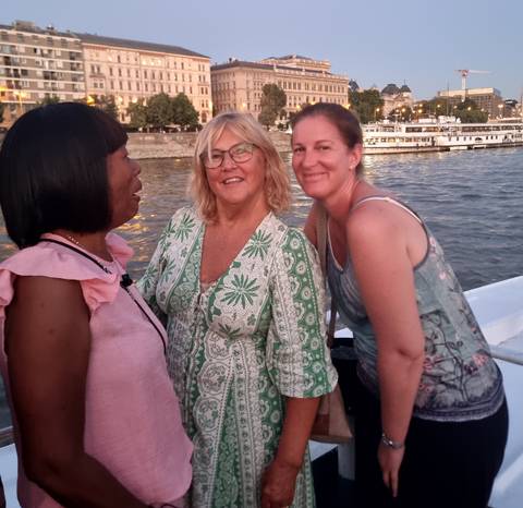 Three women chatting and smiling on a boat with a city backdrop.