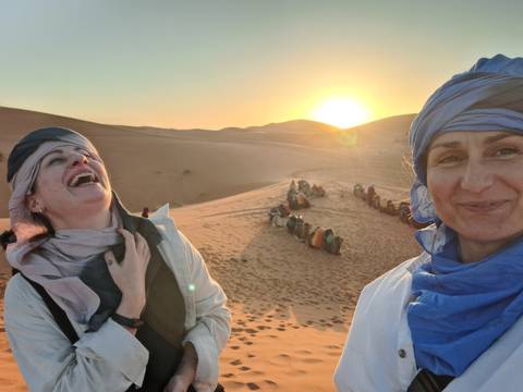       Two women laughing in the desert at sunset with a group in the background.
  
