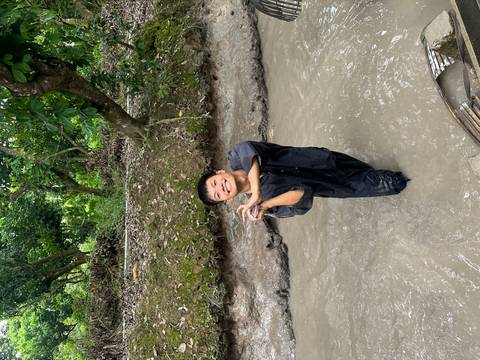 Person standing in a muddy waterway, surrounded by lush greenery.
