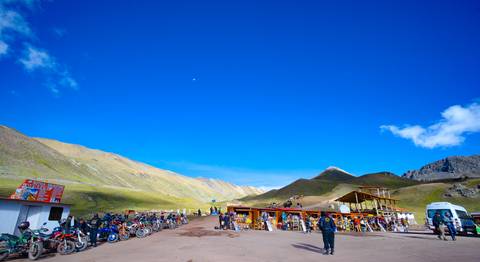 Busy open area with stalls and many motorbikes, surrounded by mountains.