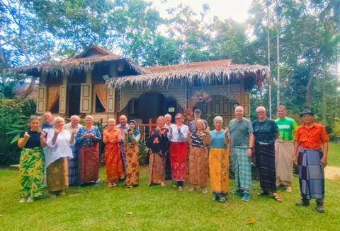       Group of people in traditional attire in front of a thatched-roof structure.
  