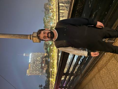       Person posing by the riverside at night in front of a lit hotel.
  