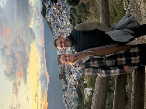      A couple posing with a view of a blue city on the hillside during sunset.
  