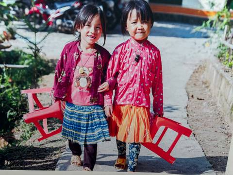 Two young girls in traditional attire carrying plastic chairs.