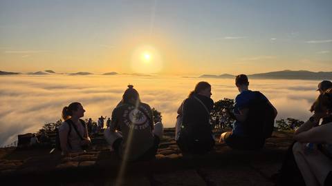       Group of people sitting on a rock enjoying a sunrise view over a sea of clouds.
  