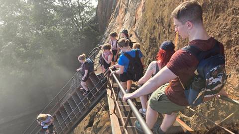       Visitors climbing a steep metal staircase attached to rock face under sunlight.
  