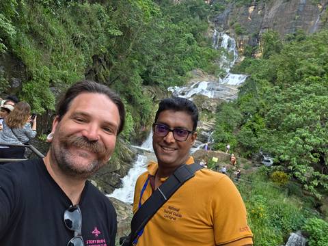 Two men taking a selfie with a waterfall and greenery in the background.