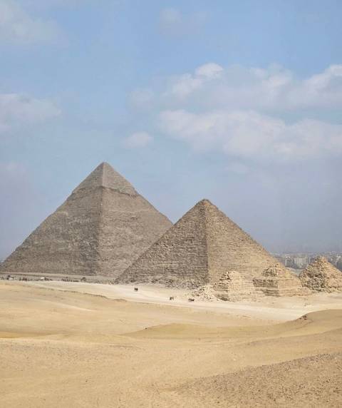       Two pyramids stand in the desert with a city view in the background.
  