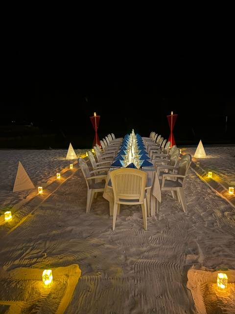       A long dinner table set up on a beach at night with glowing lights.
  