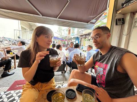 A couple drinking on a street-side cafe with pedestrians in the background.