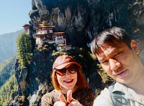       Two people smiling with a monastery perched on a cliff in the background.
  