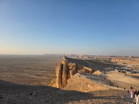 Edge of a desert cliff with cars and people overlooking the landscape.