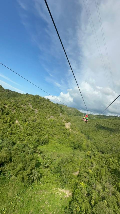 People zip-lining over lush green hills under a blue sky.