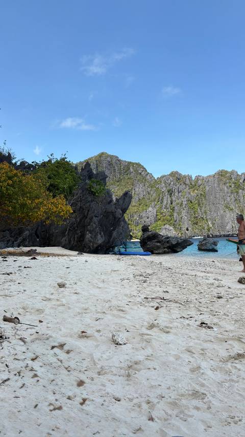 A rocky beach with a man standing, surrounded by lush green vegetation and turquoise waters.
