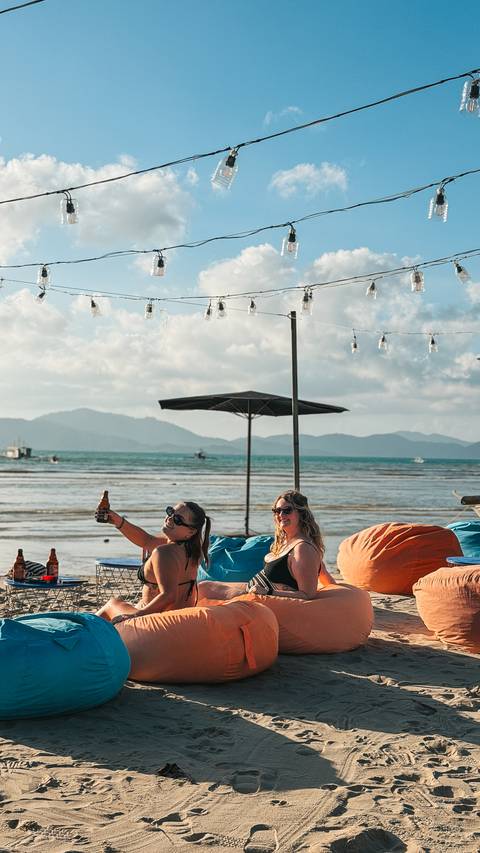       Two women enjoying drinks on a beach with mountains in the background.
  