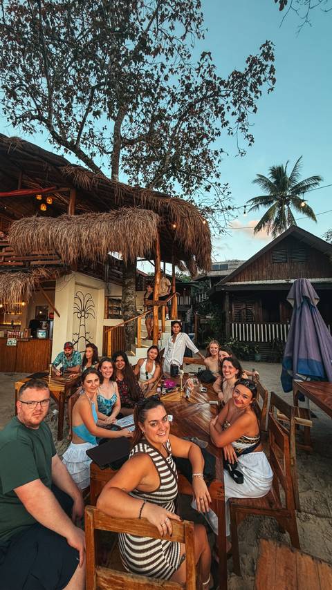       A group of people dining at a beach restaurant with a traditional roof.
  
