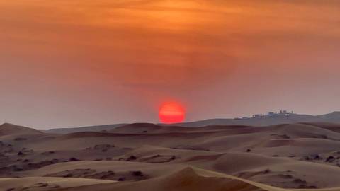       Sunset over desert dunes with a building on the horizon.
  