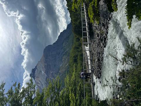       Two people crossing a bridge over a river with mountains.
  