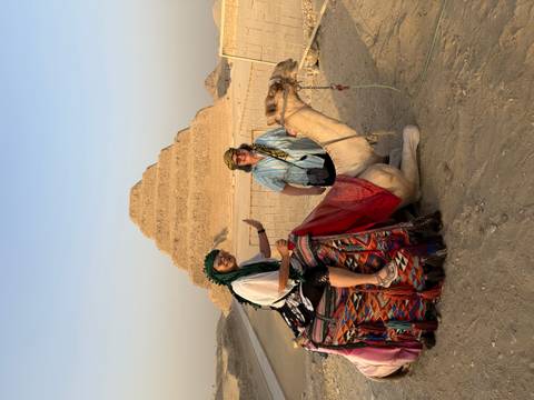 Two women on a camel in front of a pyramid.