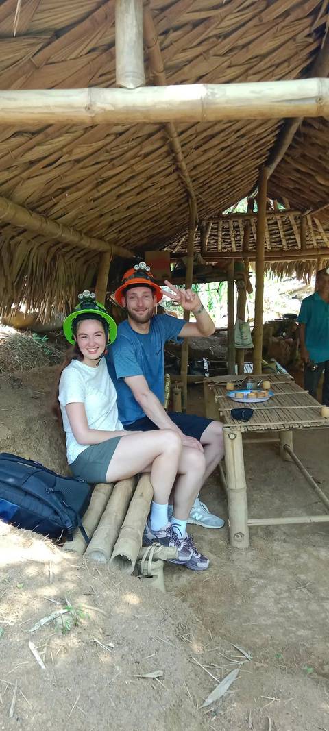       Two people sitting under a thatched roof with bamboo tables, wearing helmets and smiling.
  
