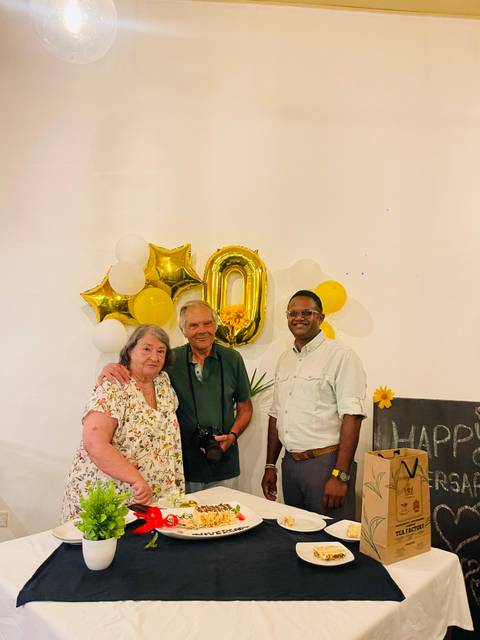 Three people standing indoors with balloons and a 'Happy' sign in the background.