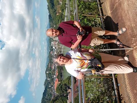 Elderly couple posing at a scenic overlook with a camera and greenery.