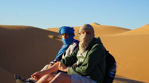       Two people sitting on desert sand dunes wearing headscarves.
  