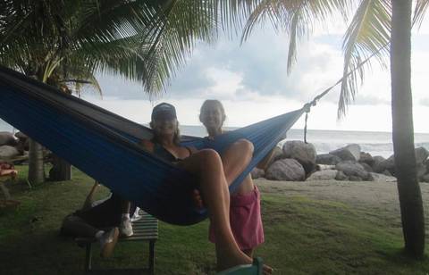       Two people relaxing in a hammock near the beach, cloudy sky, and palm trees.
  