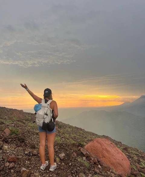 A person with a backpack gesturing towards a sunset over a mountainous landscape.