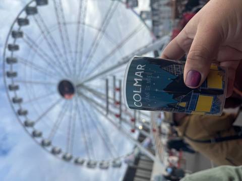 A ferris wheel in the background with a hand holding a cup labeled 'Colmar' in the foreground.