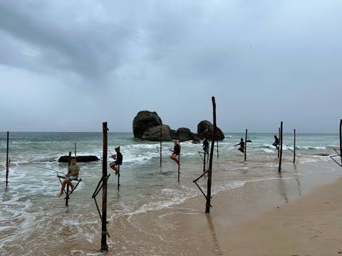 Fishermen seated on poles in the ocean.