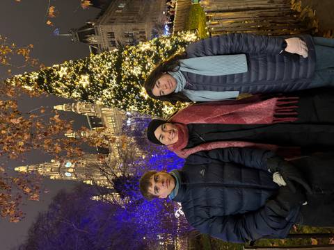       Three people posing at a Christmas market with a lit-up tree behind them.
  