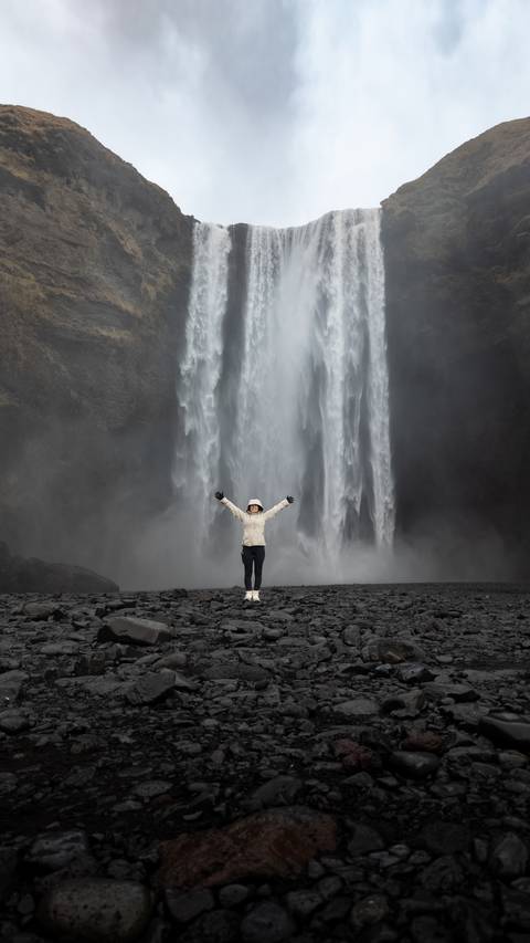       Person standing with arms raised in front of a large waterfall.
  