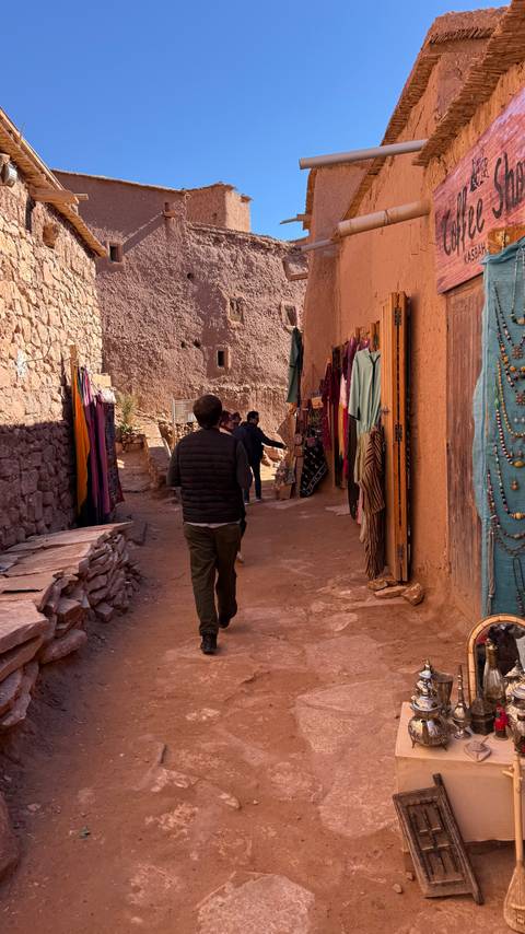       People walking through a narrow street with shops and colorful textiles.
  