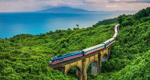       Train traveling through mountainous terrain with lush vegetation and distant view of the sea.
  