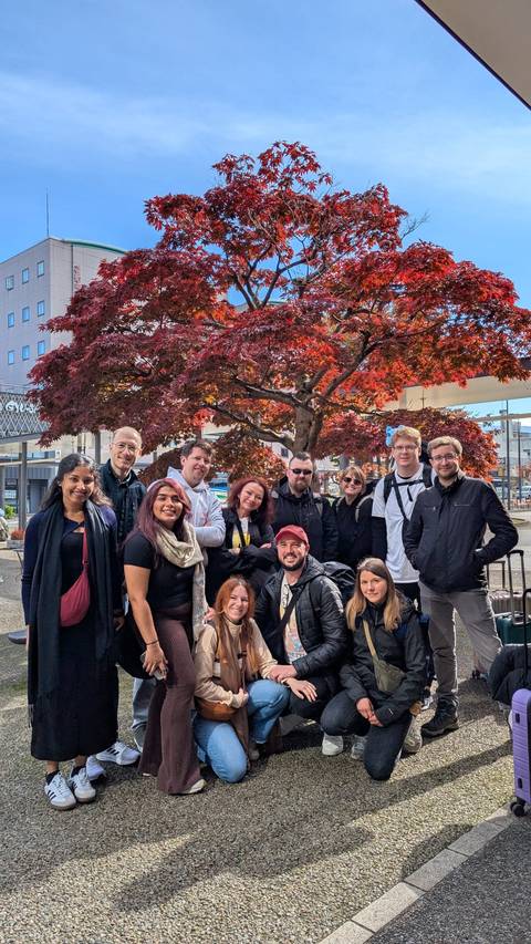 Group of people posing in front of a tree with red leaves in an urban setting.