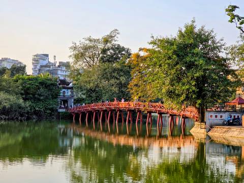 The iconic red bridge in Hanoi over a calm lake with green trees and buildings.