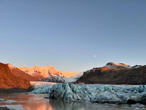 Glacier and mountains bathed in pink light with the moon overhead.