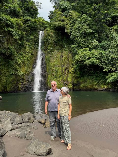 Two people standing in front of a waterfall in a forest setting.