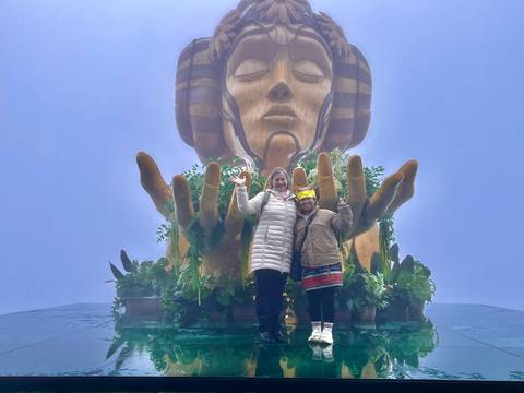 Two people posing in front of a large hand and head sculpture with vegetation.