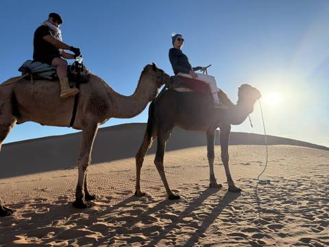 Two people riding camels in the desert during sunset.