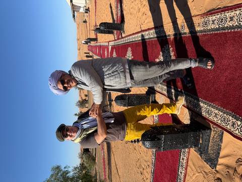Two men in traditional attire standing on a red carpet in a desert setting.