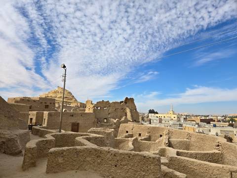       View of the Shali Fortress ruins in Siwa Oasis under a cloudy blue sky.
  
