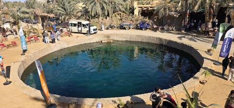 Tourists visiting a circular natural pool surrounded by palm trees at Siwa Oasis.