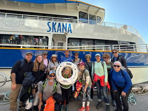       Group photo in front of a ship named 'Skana' with people smiling.
  
