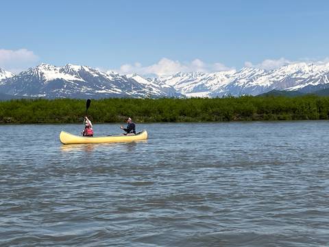       Two people canoeing on a lake with snow-capped mountains in the background.
  