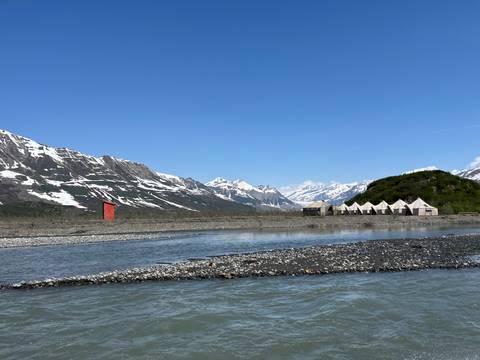       Tents with mountains and a river visible in the background under a clear blue sky.
  