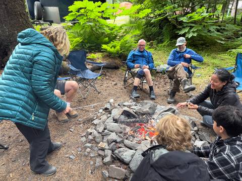       People sitting in chairs around a campfire roasting marshmallows.
  