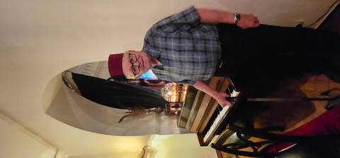 Elderly man in traditional attire standing by a piano.