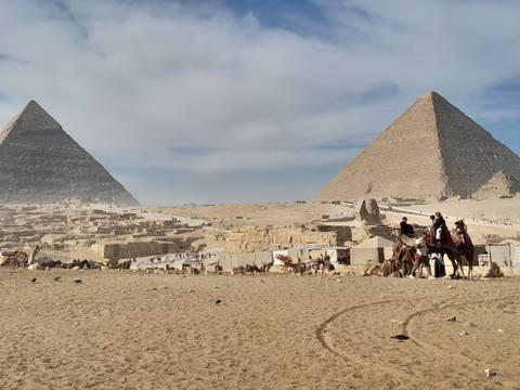       Two pyramids in the desert with camels and people.
  